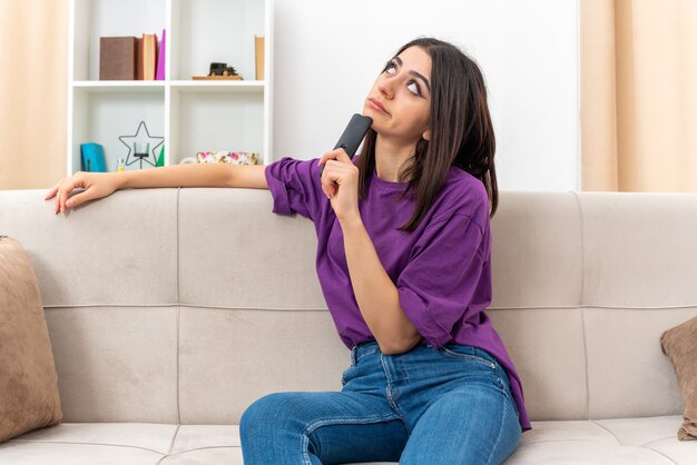 Young girl in casual clothes holding tv remote looking aside puzzled sitting on a couch in light living room