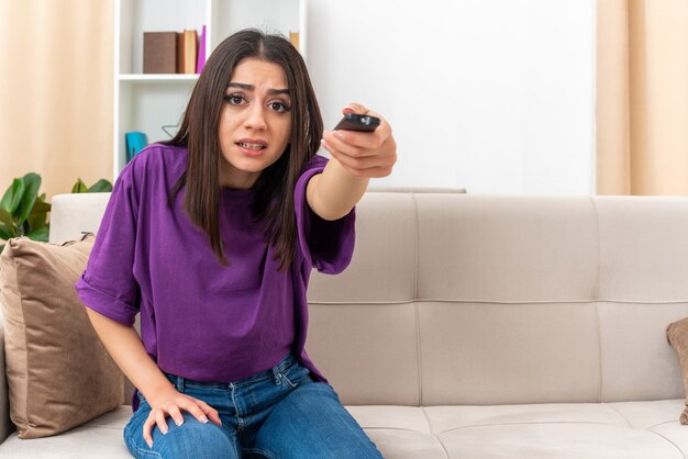 Young girl in casual clothes holding tv remote watching tv looking confused sitting on a couch in light living room