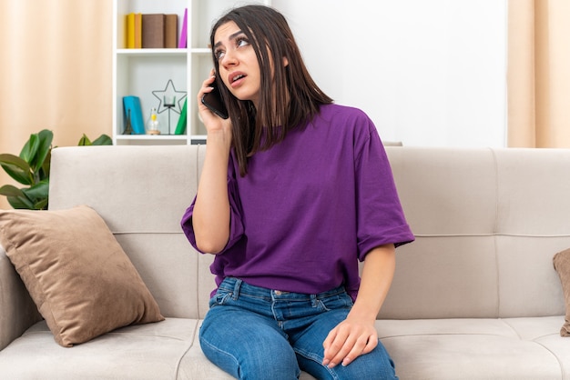 Young girl in casual clothes looking annoyed while talking on mobile phone sitting on a couch in light living room
