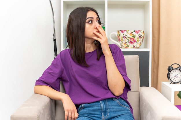 Free photo young girl in casual clothes looking aside amazed and surprised covering mouth with hand sitting on a chair in light living room