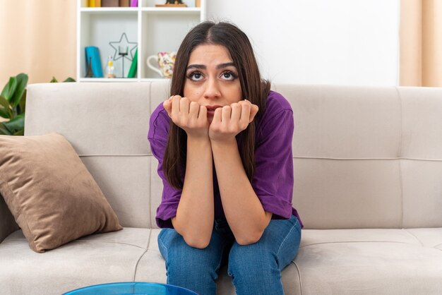 Young girl in casual clothes looking aside with hands on chin depressed with sad expression sitting on a couch in light living room