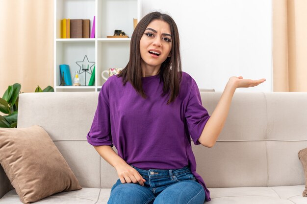 Young girl in casual clothes looking confused raising arm in displeasure and indignation sitting on a couch in light living room