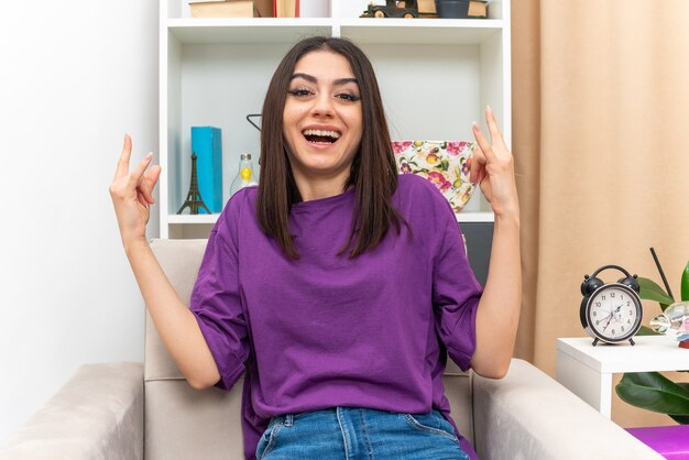 Young girl in casual clothes looking happy and excited making rock symbol sitting on a chair in light living room