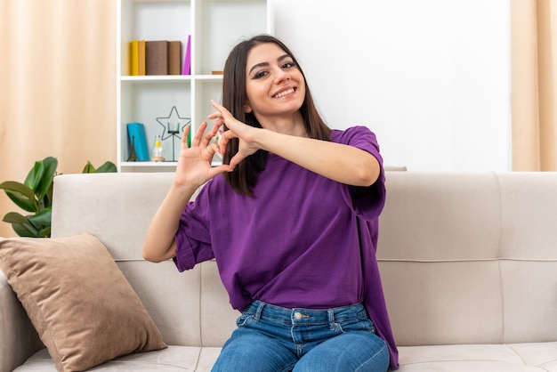 Young girl in casual clothes looking happy and positive smiling cheerfully making heart gesture with fingers sitting on a couch in light living room