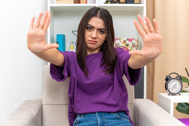 Young girl in casual clothes looking with serious face making stop gesture with hands sitting on a chair in light living room