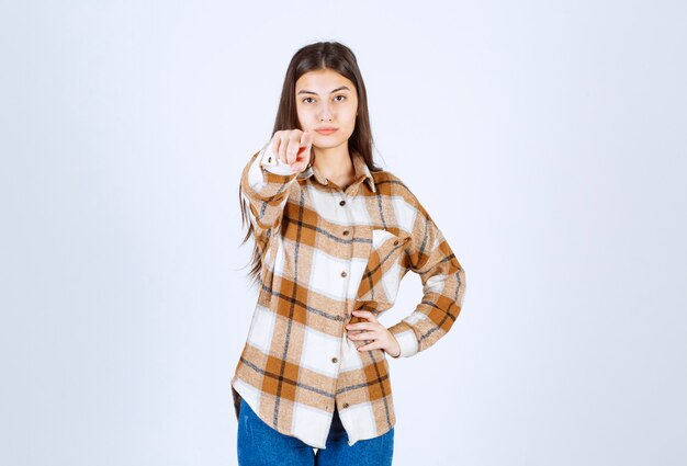 Young girl in casual clothes standing and pointing at camera on white wall. 