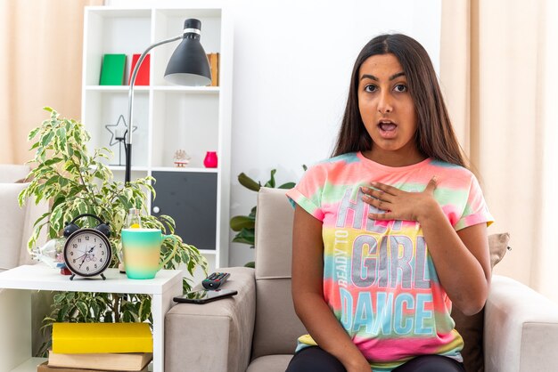 Young girl in casual clothes  surprised pointing at herself sitting on the chair in light living room