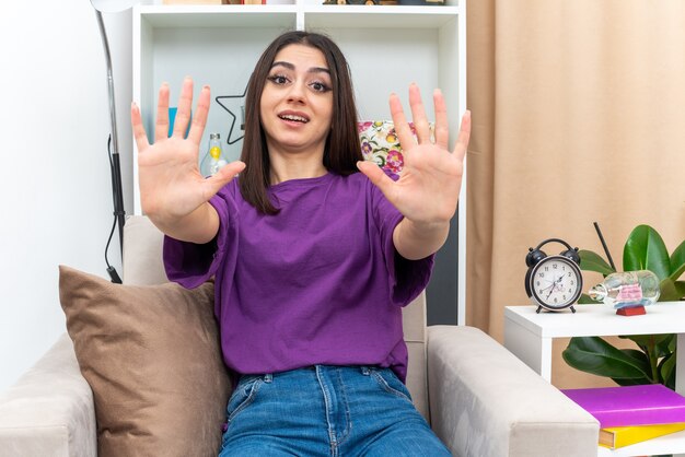 Young girl in casual clothes  worried holding hands out as telling do not come closer sitting on a chair in light living room