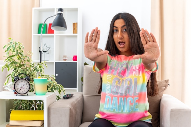 Young girl in casual clothes  worried making stop gesture with hands sitting on the chair in light living room