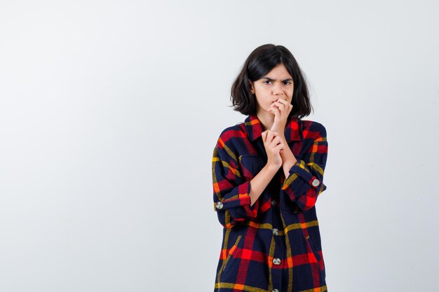 Young girl in checked shirt biting fingers and looking excited , front view.