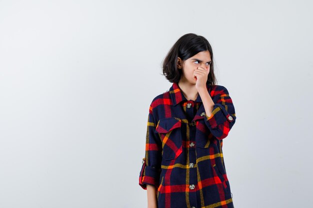 Young girl in checked shirt covering mouth and nose with hand, looking away and looking exhausted , front view.