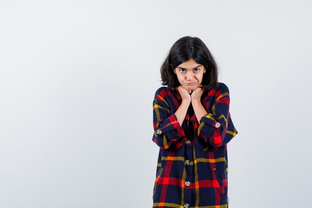 Young girl in checked shirt holding hands under chin and looking cute , front view.