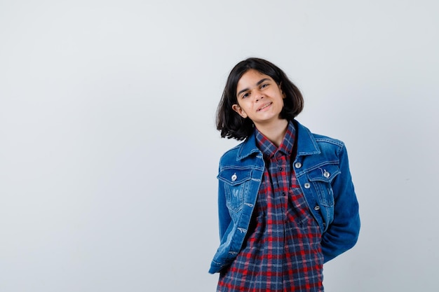 Foto gratuita giovane ragazza in camicia a quadri e giacca di jeans che si tiene per mano dietro la vita e sembra carina, vista frontale.