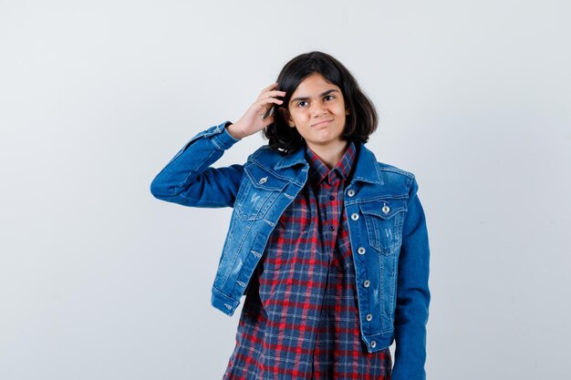 Young girl in checked shirt and jean jacket scratching head and looking happy , front view.