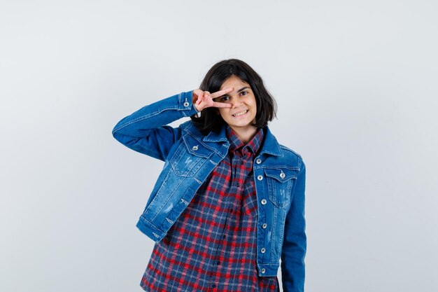 Young girl in checked shirt and jean jacket showing v sign on eye and looking pretty , front view.