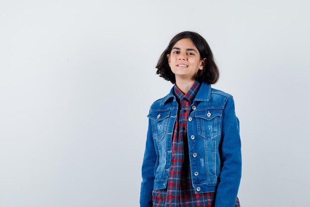 Young girl in checked shirt and jean jacket standing straight and posing at camera and looking happy , front view.