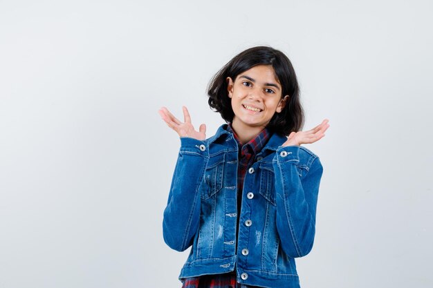 Young girl in checked shirt and jean jacket stretching hands in questioning manner and looking pretty , front view.