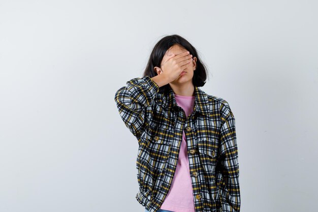 Young girl in checked shirt and pink t-shirt covering eyes with hand and looking timid, front view.