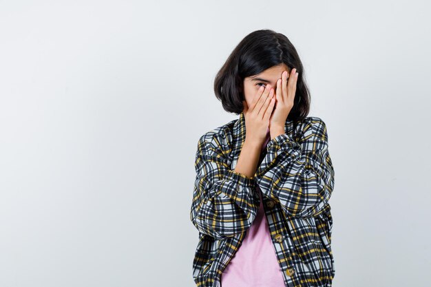 チェックシャツとピンクのTシャツを着た少女が顔を手で覆い、臆病に見える