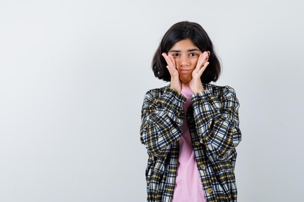 チェックシャツとピンクのTシャツを着た少女が顔に手をつないで、臆病に見える、正面図。