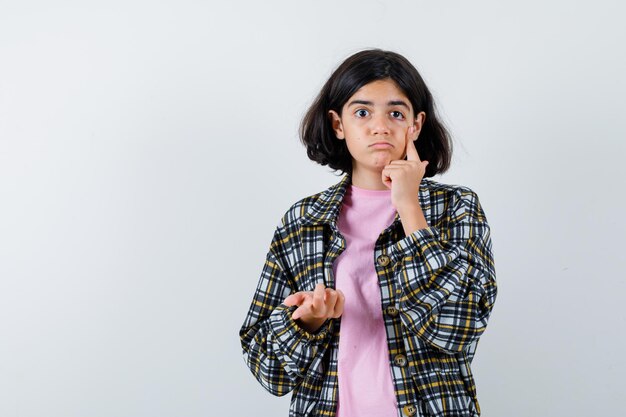 チェックシャツとピンクのTシャツの若い女の子は、目の領域に人差し指を置き、拳を握りしめ、きれいに見える、正面図。