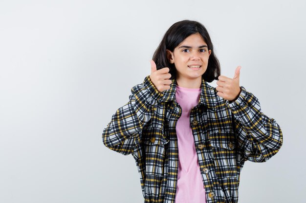 Young girl in checked shirt and pink t-shirt showing thumbs up with both hands and looking happy , front view.