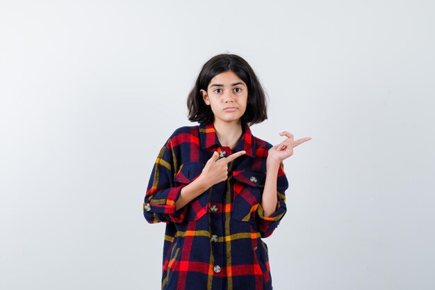 Young girl in checked shirt pointing right with index fingers and looking pretty , front view.