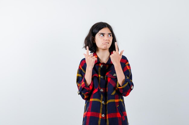 Young girl in checked shirt pointing up with index fingers and looking serious , front view.