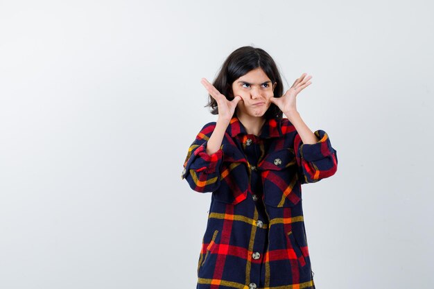 Young girl in checked shirt pulling cheeks with fingers, looking upward and looking pensive , front view.