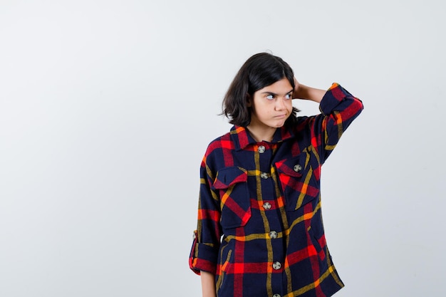 Young girl in checked shirt putting hand on head, looking away and looking pensive , front view.
