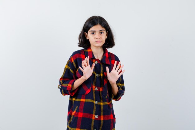 Young girl in checked shirt raising hands in surrender pose and looking scared , front view.