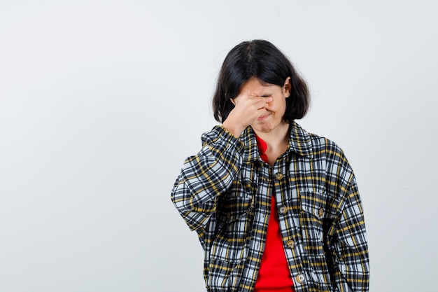 Young girl in checked shirt and red t-shirt covering face with hand and looking serious , front view.