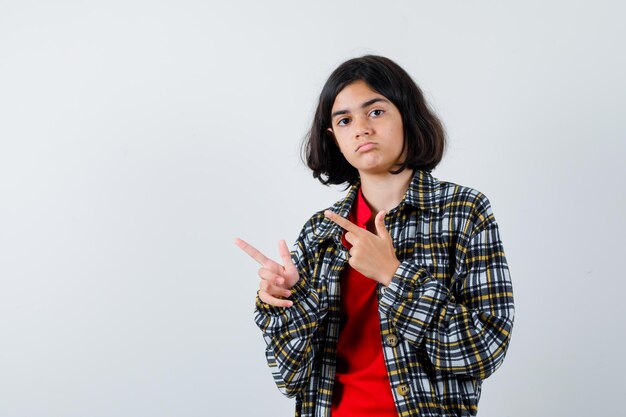 Young girl in checked shirt and red t-shirt pointing left with index fingers and looking serious , front view.