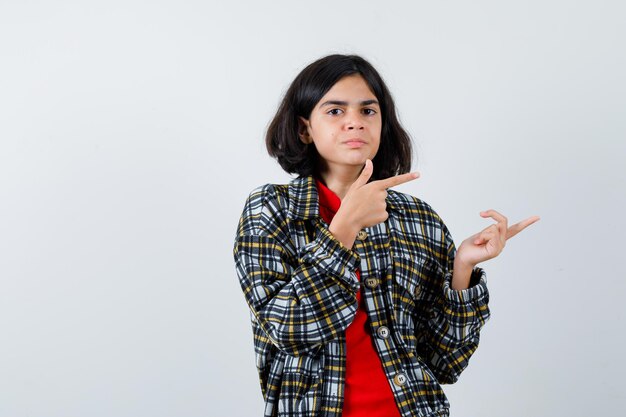 Young girl in checked shirt and red t-shirt pointing right with index fingers and looking serious , front view.