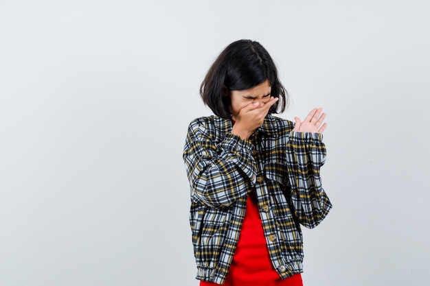 Young girl in checked shirt and red t-shirt putting hand on mouth, sneezing and looking serious , front view.