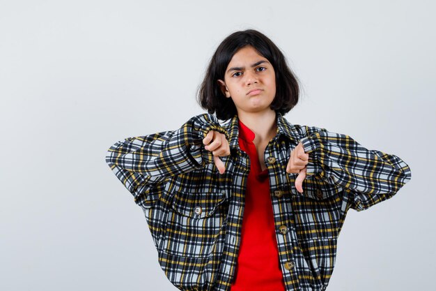 チェックのシャツと赤いTシャツを着た少女が両手で親指を下に向けて、不機嫌そうに見える、正面図。