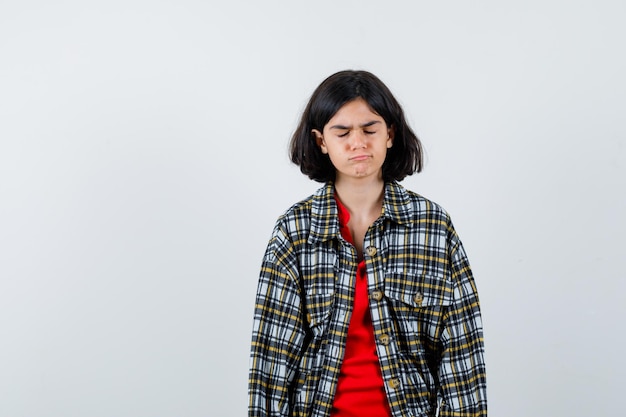 Giovane ragazza in camicia a quadri e t-shirt rossa in piedi dritta, chiudendo gli occhi e posando alla telecamera e guardando seria, vista frontale.