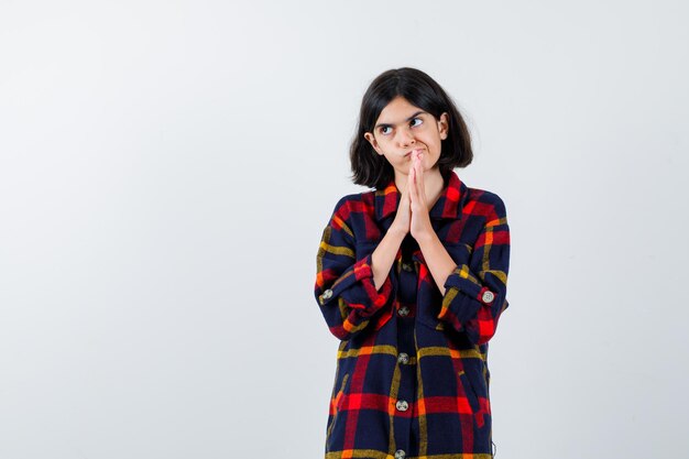 Young girl in checked shirt showing namaste gesture, looking away and looking pensive , front view.