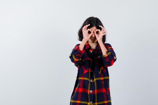 Young girl in checked shirt showing ok signs on eyes and looking cute , front view.