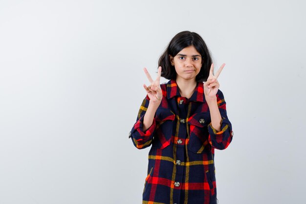 Young girl in checked shirt showing peace gestures and looking cute , front view.