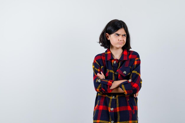 Young girl in checked shirt standing arms crossed, looking away and looking harried , front view.