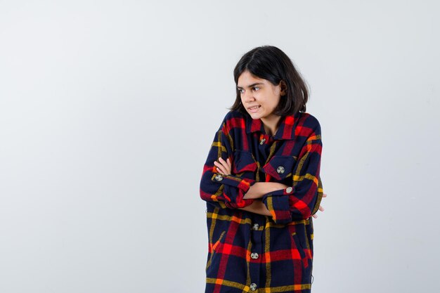 Young girl in checked shirt standing arms crossed, looking away and looking timid , front view.