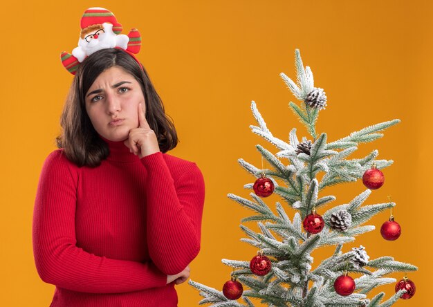Молодая девушка в рождественском свитере с забавной повязкой на голову рядом с елкой на оранжевом фоне
