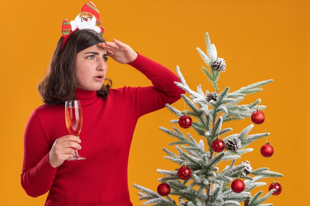 Young girl in christmas sweater wearing funny headband holding glass of champagne looking far away with hand over head standing next to a christmas tree over orange wall