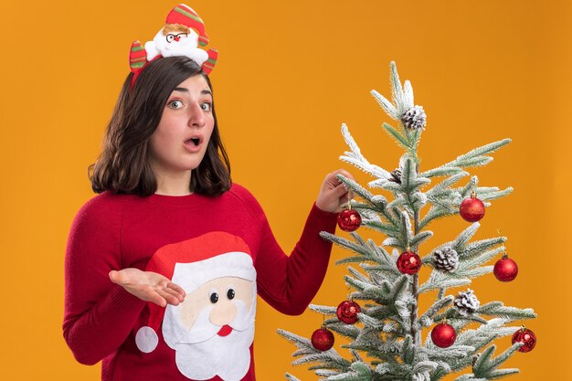 Young girl in christmas sweater wearing funny headband looking at camera confused standing next to a christmas tree over orange background