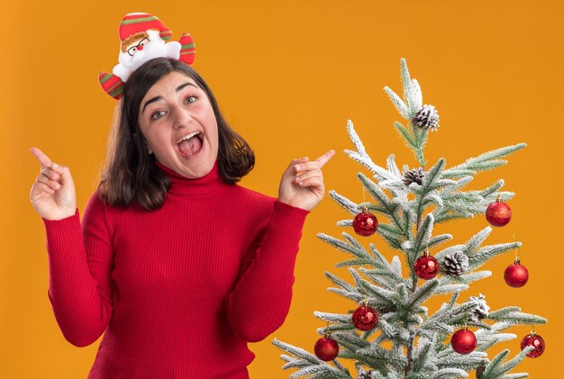 Young girl in christmas sweater wearing funny headband looking at camera happy and excited next to a christmas tree over orange background