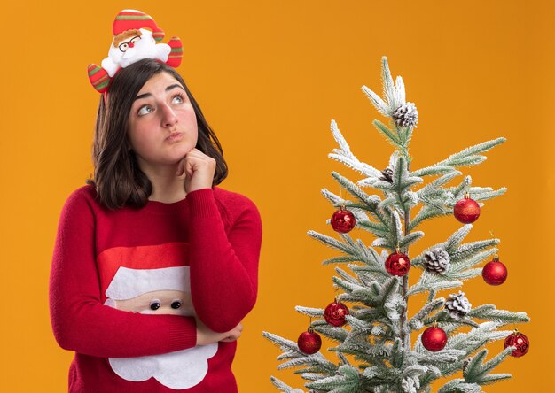 オレンジ色の壁の上のクリスマスツリーの横に立って困惑して見上げる面白いヘッドバンドを身に着けているクリスマスセーターの少女