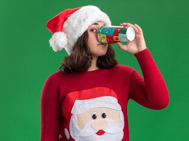 녹색 벽 위에 서있는 다채로운 종이 컵에서 마시는 산타 모자를 쓰고 크리스마스 스웨터에 어린 소녀