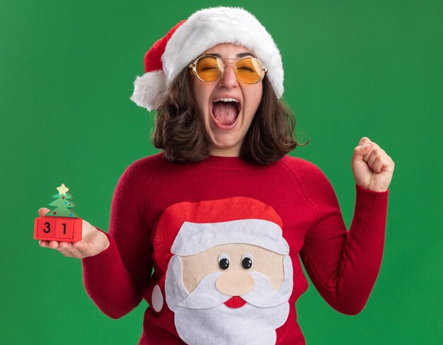 サンタの帽子をかぶったクリスマスセーターの少女と新年の日付のおもちゃの立方体を保持しているメガネ緑の壁の上に立っているクレイジー幸せで興奮した握りこぶし
