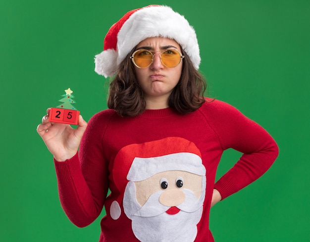 Young girl in christmas sweater wearing santa hat and glasses holding toy cubes with number twenty five  displeased pursing lips standing over green wall